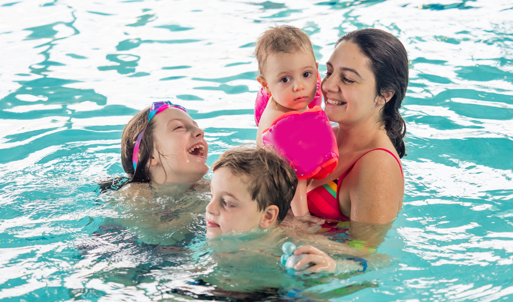 Photo of mother and three children of mixed ages, smiling and interacting together in the swimming pool, with the youngest child in her arms.